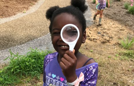 Little girl holds a magnifying glass to her eye while smiling at the camera