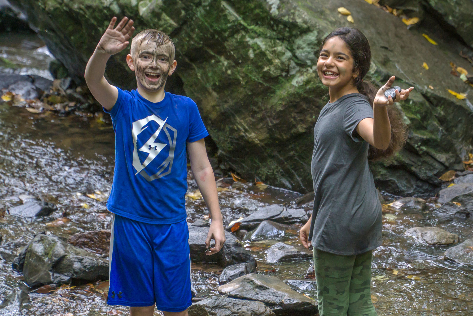 Two school children wave at the camera while playing in water at the base of a waterfall in the Smokies