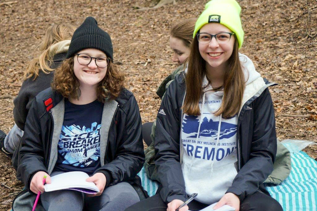 Two students pause from writing observations in nature journals to smile at the camera