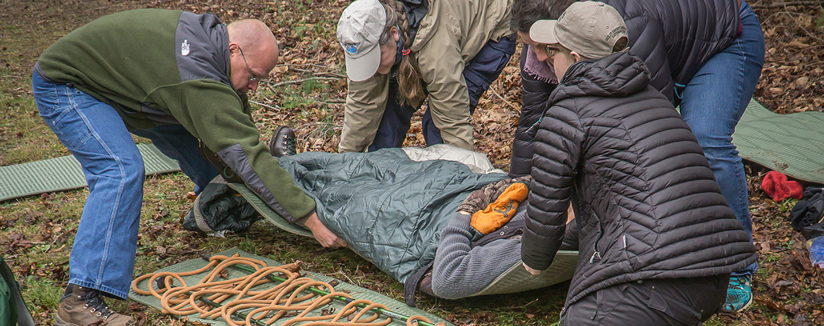 A group of people practice moving someone onto a stretch in a mock emergency situation outdoors.