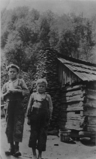 In a black and white historical photo, two boys stand in front of a log cabin.